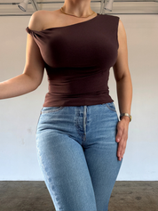 Person wearing a brown one-shoulder top and blue jeans against a plain background