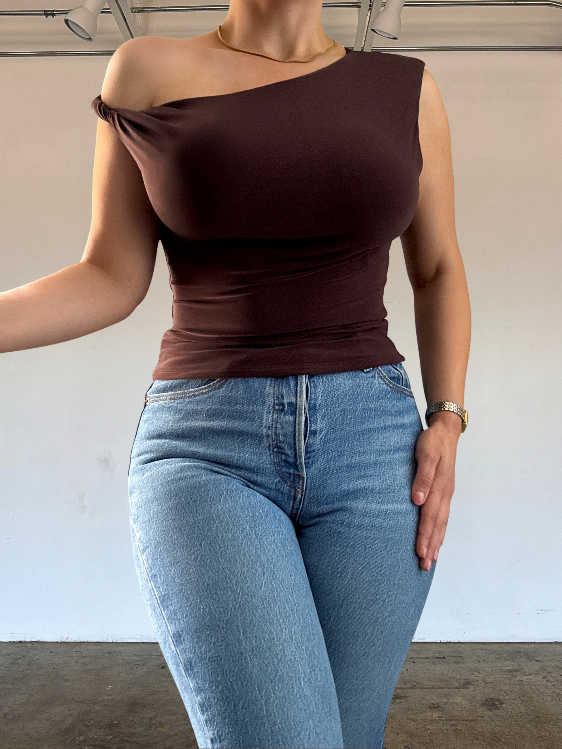 Person wearing a brown one-shoulder top and blue jeans against a plain background